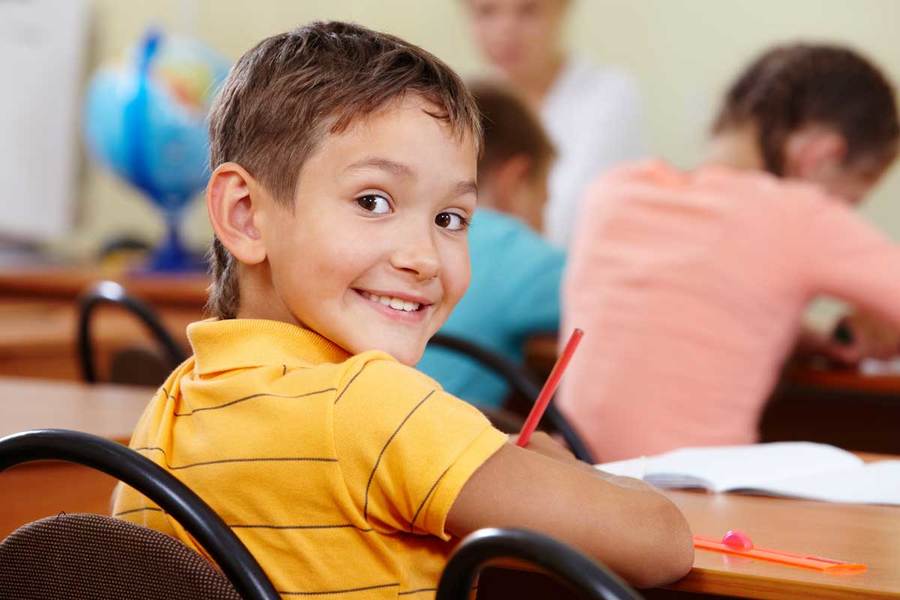 A smiling student diagnosed with ADHD turns around in his chair while working on an assignment in a classroom setting.