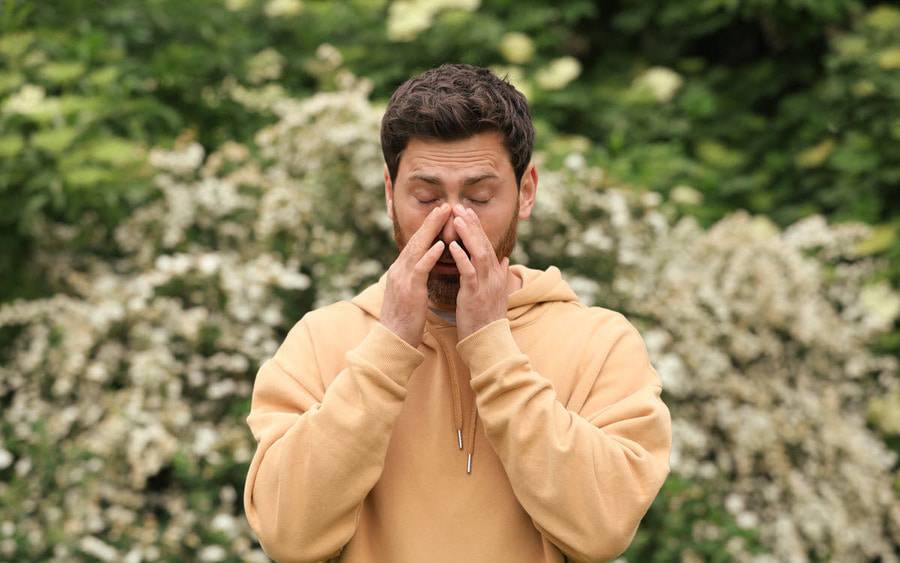 A young man who is standing outdoors with plants in bloom in the background lightly touches his nose with both hands with his eyes closed.