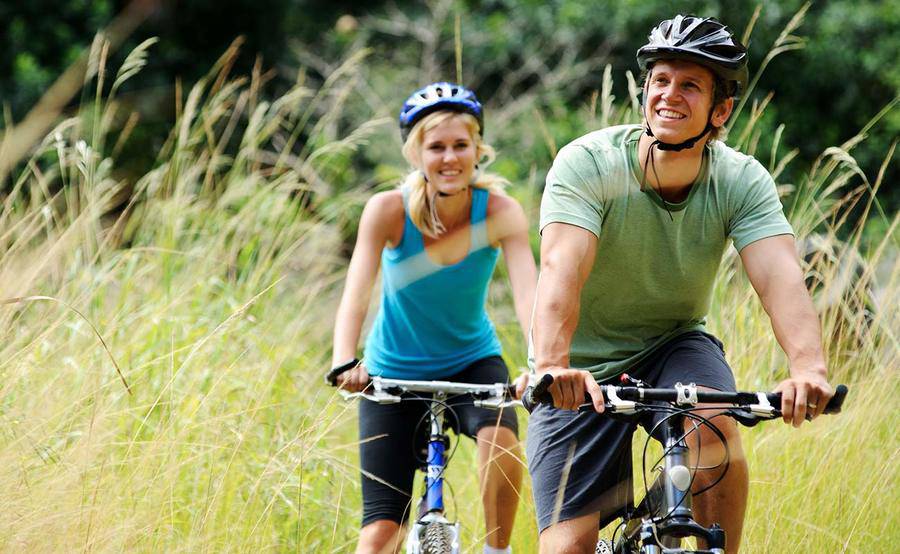 A woman and man riding bicycles together in tall grass.