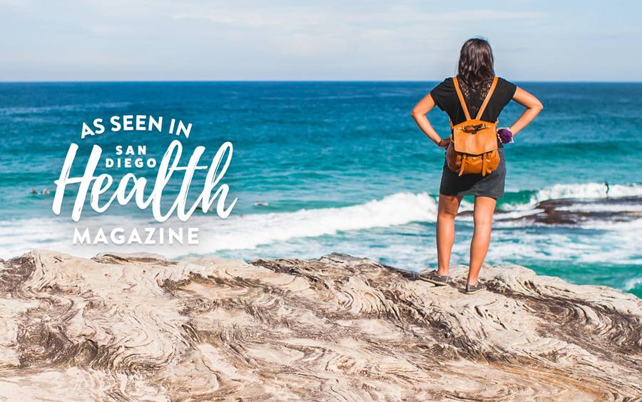 A woman stands on an ocean bluff looking out over the water. San Diego Health Magazine