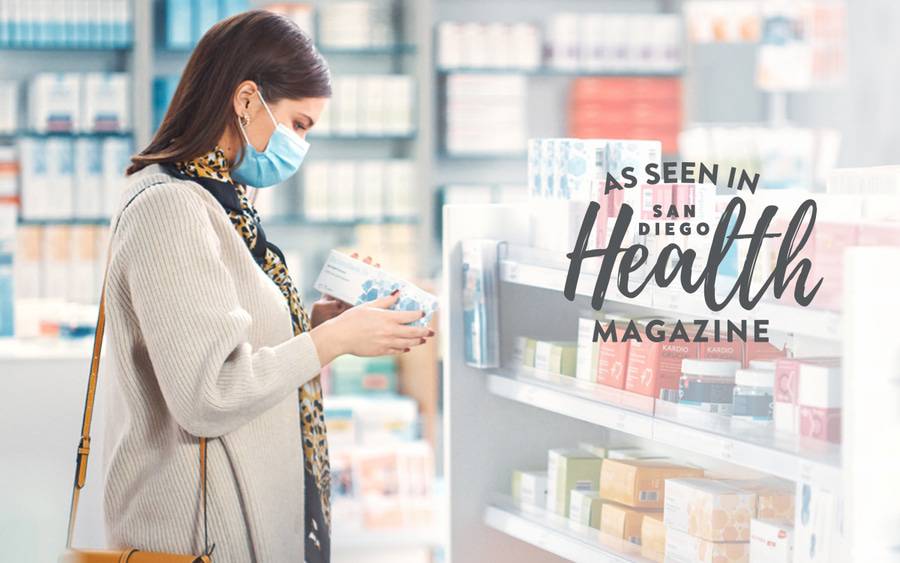 A woman wearing a surgical facemask to prevent getting sick reads the package of a natural immunity product in a pharmacy store - SD Health Magazine.