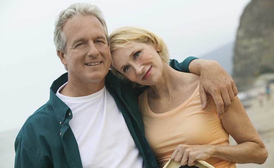 A smiling middle-aged Caucasian couple on the beach represents the full life that can be led with the latest chemotherapy techniques that maximize comfort.
