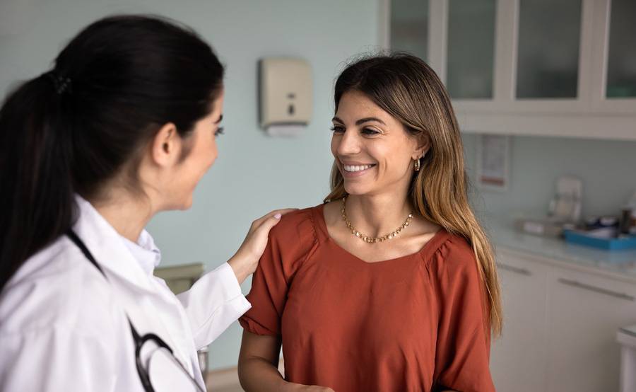 A compassionate female doctor is putting her hand on the shoulder of her female patient and they are smiling at each other.