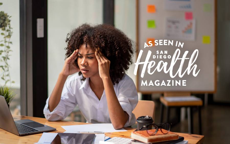A young woman sits at a desk and holds her hands on the sides of her head as she looks concerned looking at a laptop computer. San Diego Health Magazine