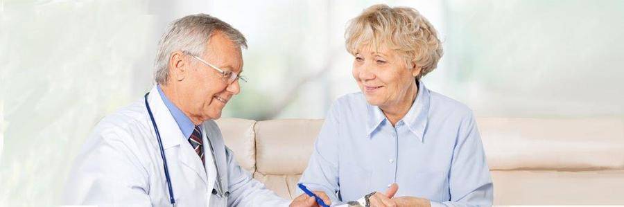 A relaxed elderly woman talks with her physician while seated on a couch in an indoor clinical setting.