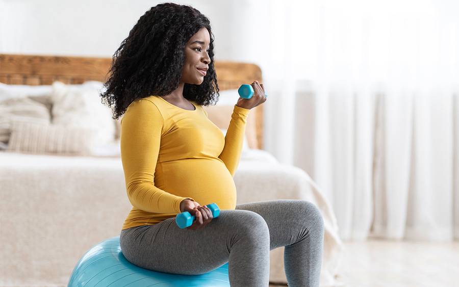 A pregnant woman sits in her bedroom exercising with light weights.
