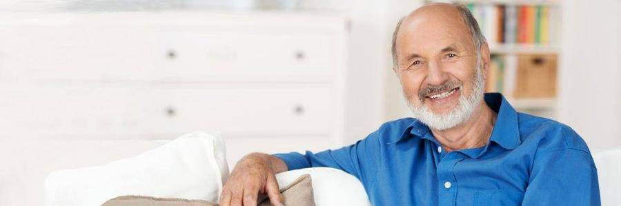 A smiling elderly gentleman relaxes on a white couch in a pleasant indoor setting.