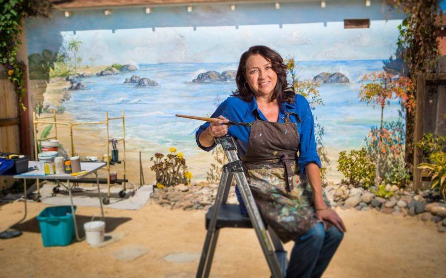 Mural artist, Gloria Favela, sits on a step ladder in a paint-splattered apron with a paint brush in her hand.