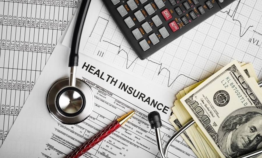 A stack of money, pen, calculator and stethoscope lie on health insurance forms depicting disruption in healthcare.