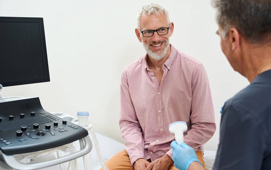 A male patient with gray hair smiles as the provider gets ready to use an ultrasound to monitor inflammatory bowel disease.
