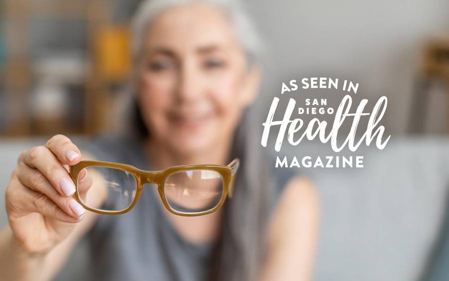 A woman with gray hair is blurred behind her eyeglasses that she's holding in front of her. San Diego Health Magazine