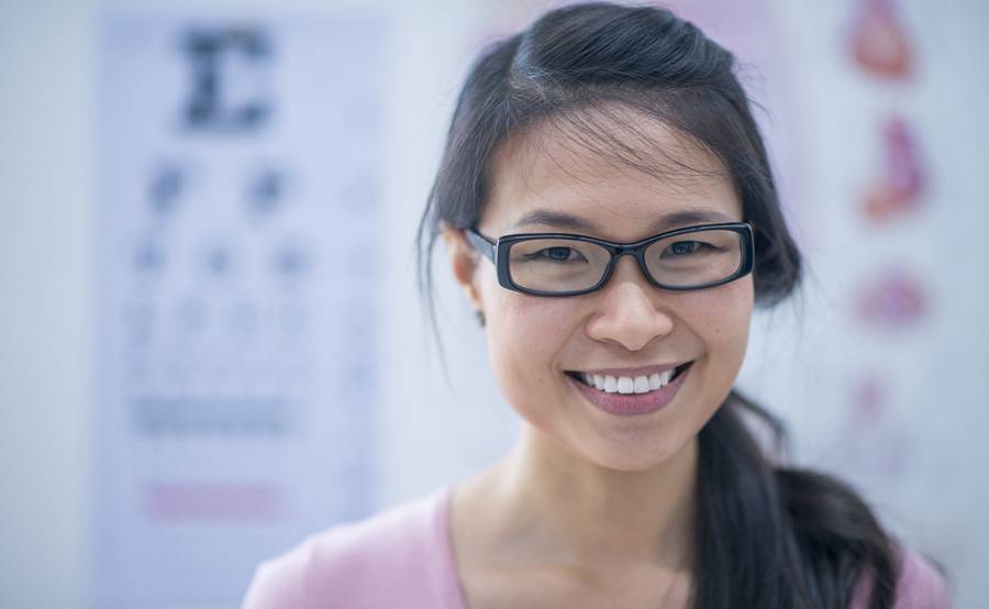 A young woman with glasses smiles in front of an eye chart, representing how you can see your best with ophthalmology and optometry care from Scripps in San Diego.