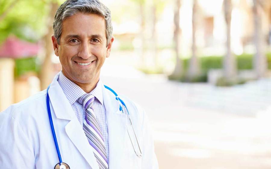 A physician stands in an outdoor setting with a white lab coat and blue stethoscope.