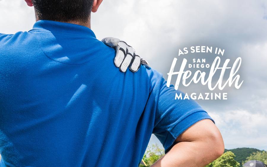 A man with a blue shirt touches his right shoulder with his left hand while he looks out toward the golf course. SD Health Magazine