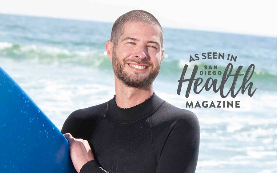 Robert Elliott, DO, an internal medicine physician at Scripps Coastal Medical Center Hillcrest, pictured holding a surfboard at the ocean in San Diego. San Diego Health Magazine