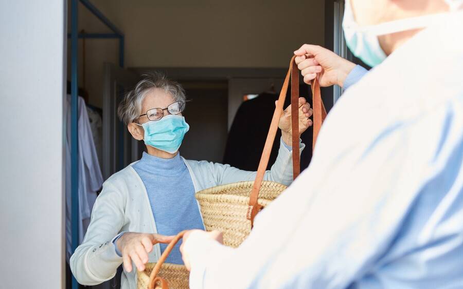 A caregiver wearing mask gives senior also wearing a mask a basket of food during coronavirus pandemic.