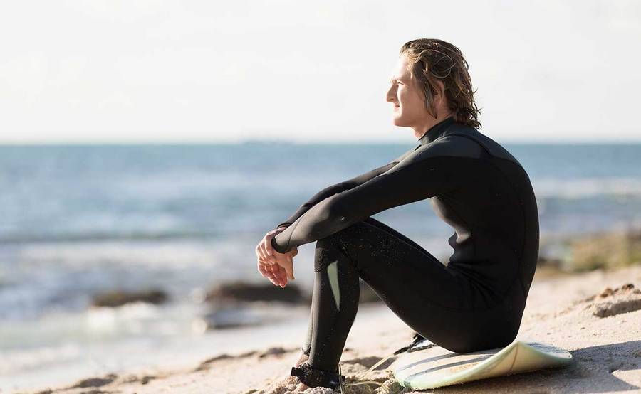 A young surfer sitting on the beach represents the full life that can be led after testicular cancer treatment.