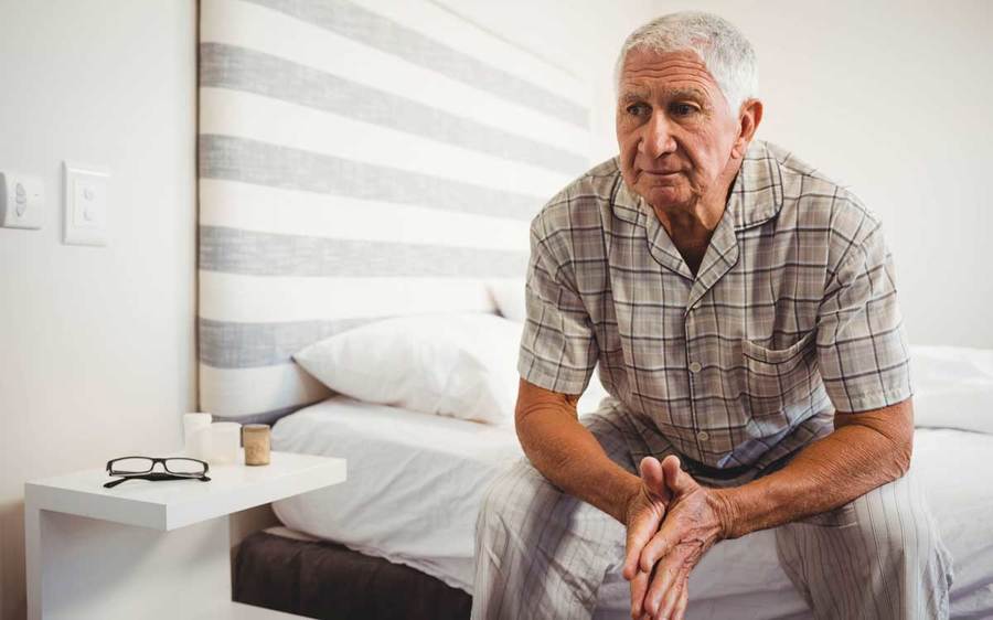 A thoughtful elderly man sits on his bed near a nightstand holding his glasses and candles.