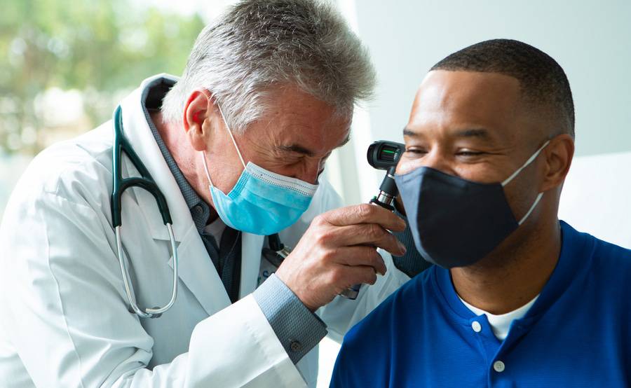 Physician performing an ear examination, with a face mask, on a patient also wearing a mask.
