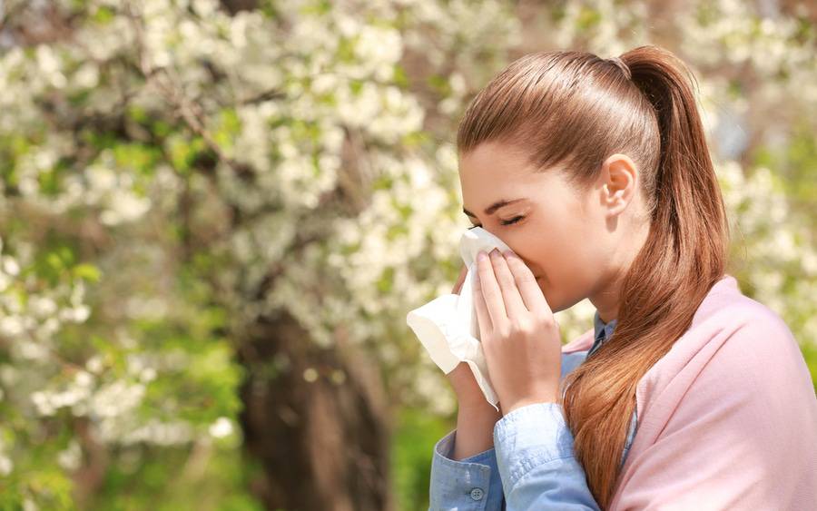 Woman sitting outside during the Spring with blooming trees while sneezing, illustrating someone suffering from allergies.