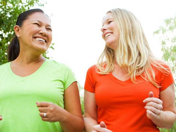 A Latina woman and a white woman smile as they enjoy a morning walking in the park for exercise.