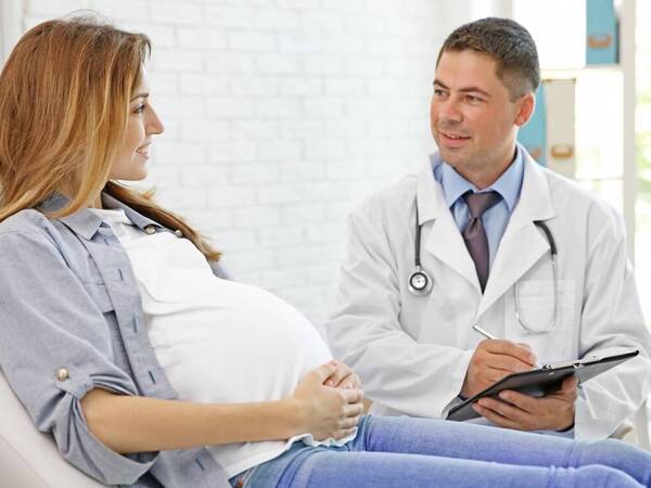 A pregnant woman sits on a bed talking to her gynecologist. Photo is used to illustrate a web post on a study that found no link between taking biologics and pregnancy complications.