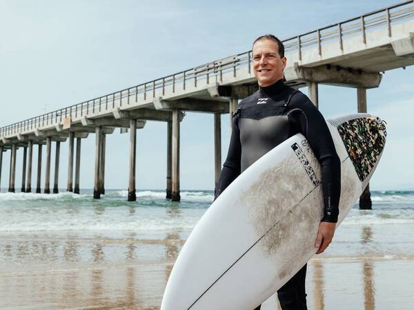 Scripps orthopedic surgeon Dr. Timothy Peppers stands on the beach with his surfboard, inspiring patients of all ages to stay active and maintain mobility.