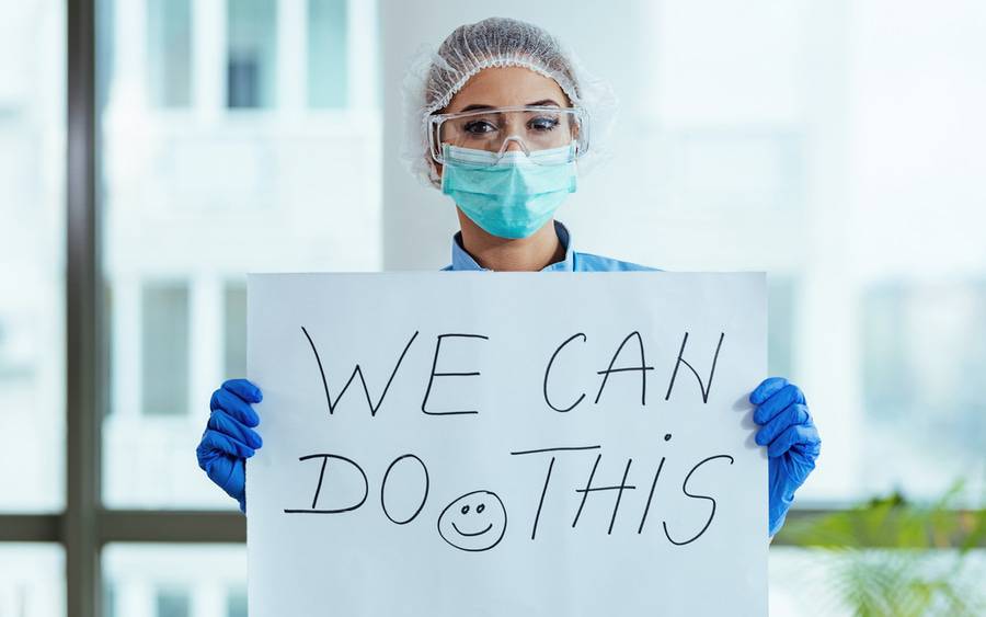 Nurse in scrubs and protective equipment holds sign saying: We Can Do This.
