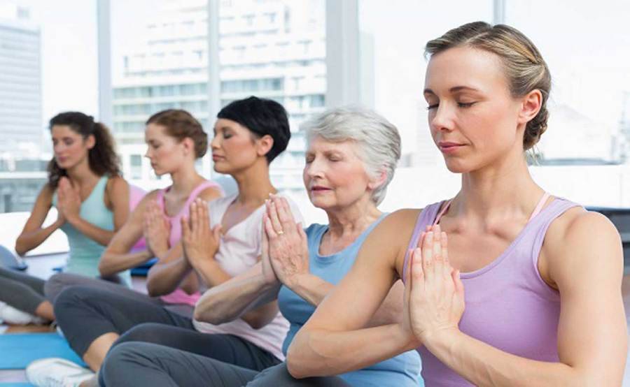 Five women of various ages seated in a yogic lotus pose with their eyes closed and palms pressed together, and city skyline in the background.