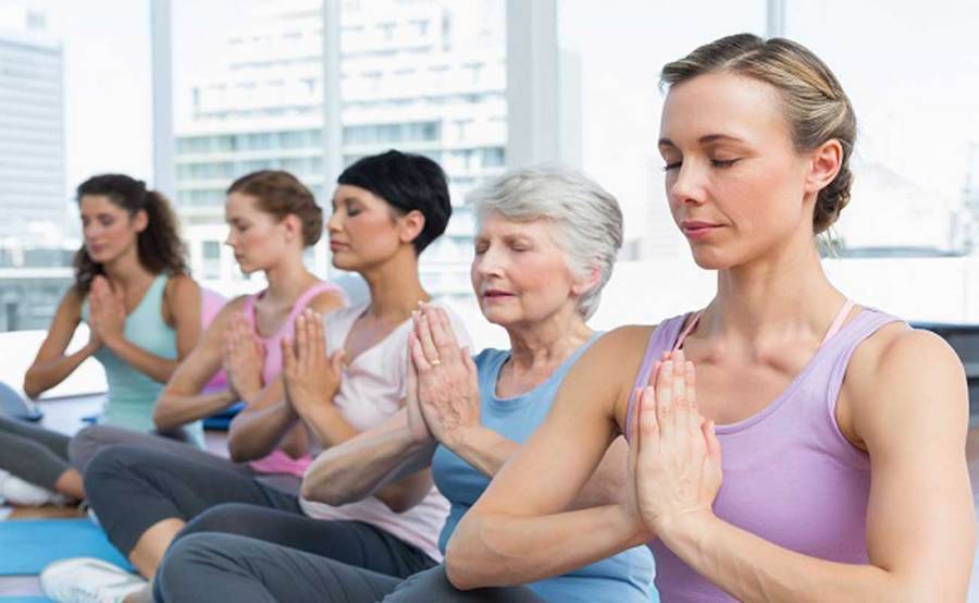 Five women of various ages seated in a yogic lotus pose with their eyes closed and palms pressed together, and  city skyline in the background.