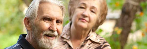 Man with gray beard smiling and serious woman wearing collared shirt in the background with green trees.