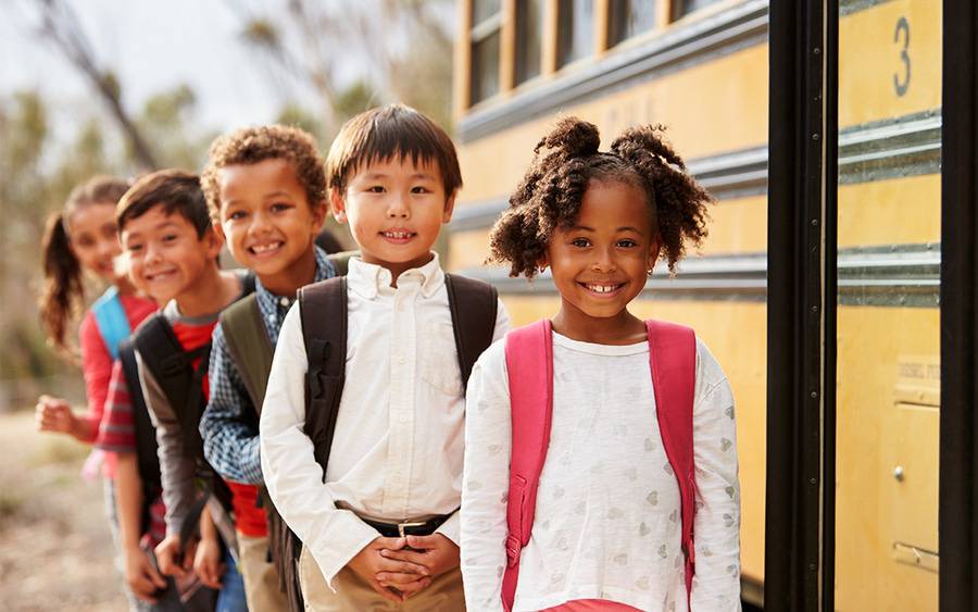 A diverse group of kids smile as they prepare to board a school bus for the first day of school.