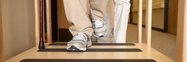 A patient wearing pants and sneakers uses a cane to practice balance with the assistance of a physical rehabilitation professional.