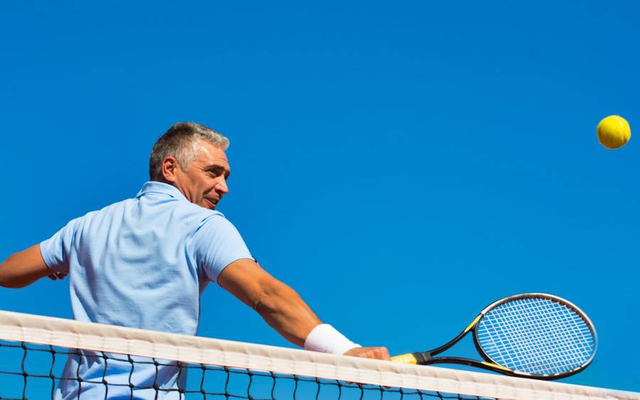 A man with gray hair is back on the tennis court as he hits a tennis ball after shoulder surgery using a new minimally invasive procedure fixes torn rotator cuffs - SD Health Magazine