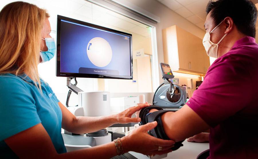 A physical therapist supports a patient's arm in a brace while looking at a monitor during a session for brain injury rehab.