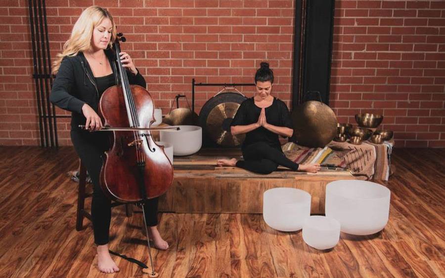 A woman plays a cello while another woman meditates near singing bowls.