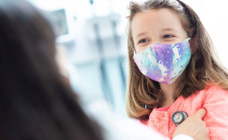 A physican performing a wellness check-up on a young girl who is wearing a unicorn printed face mask.