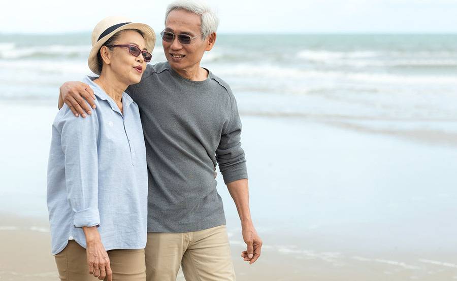 A smiling mature Asian couple walk on the beach and represent the full life that can be led after rectal cancer treatment.