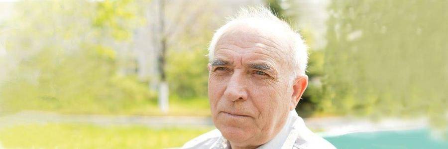 A serious-looking elderly gentleman pauses while walking in a tree-filled park.