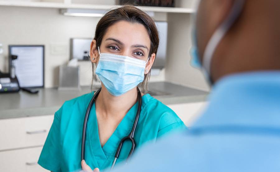 A nurse smiles behind a surgical mask while talking with a patient in the back office of a clinic.