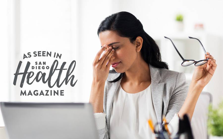 A professional woman sitting at a desk holding her head in one hand and her glasses in the other