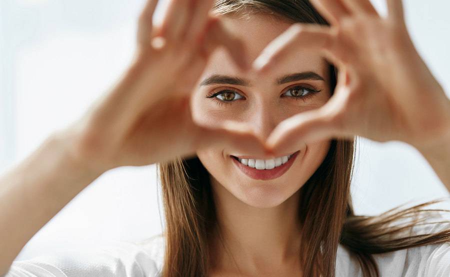 A young woman looks through her hands that are held up in the shape of a heart, representing the naturally beautiful appearance after cosmetic eye surgery.