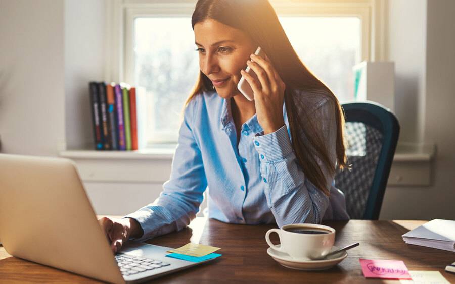 A woman working on her computer from home, while on her mobile phone.