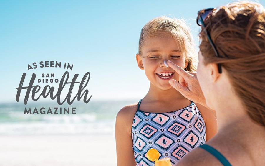 A mother putting sunscreen on her daughter's face as her daughter is laughing, illustrating sun protection during the Summer months.