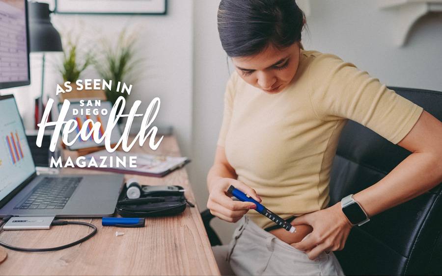 A young woman injects a weekly insulin injection into the left side of her abdomen while sitting at the computer - SD Health Magazine