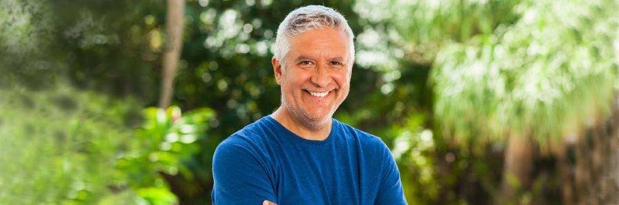 A happy middle-aged man stands with folded arms in a sunny outdoor setting with palm trees and ferns in the background.