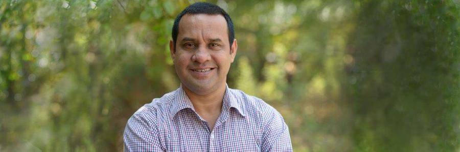 A relaxed middle-aged man wearing a checked shirt stands in a pleasant outdoor setting with greenery visible in the background.