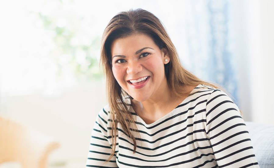 A woman smiling broadly while sitting on a sofa represents the confidence you can gain from a medically supervised weight loss program.