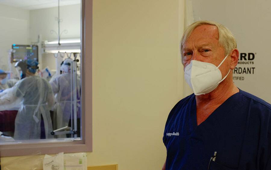 Dr. Dennis Amundson wears a medical mask while standing next to a window into an ICU room, where medical staff in PPE attend to a COVID patient.