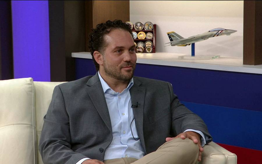 Dr. Kelley is sitting on a white couch in a newsroom with an Air Force model plane in the background on a shelf.